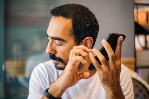 Un homme qui a les yeux fermés et qui utilise un smartphone du bout des doigts en l'ayant placé de son oreille gauche. 