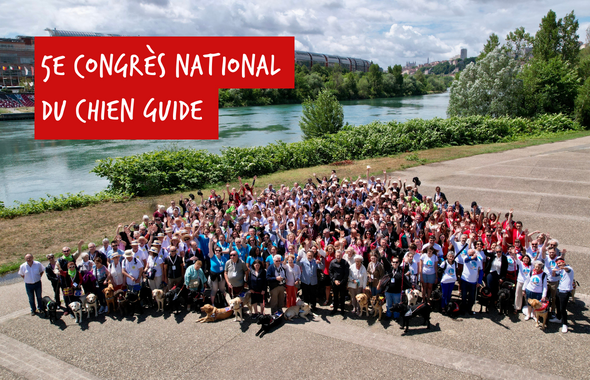 Photo du groupe de participants au Congrès National du Chien Guide 2022 à Lyon. Les personnes sont debout, sur un parvis. Derrière elles passent le Rhône et en arrière-plan se situe le Centre de Congrès. Le temps est ensoleillé.